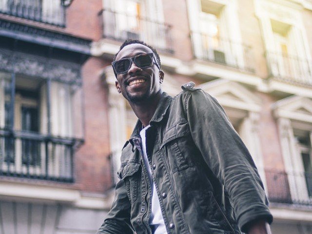 Man smiling with sunglasses on and rows of apartments behind him.