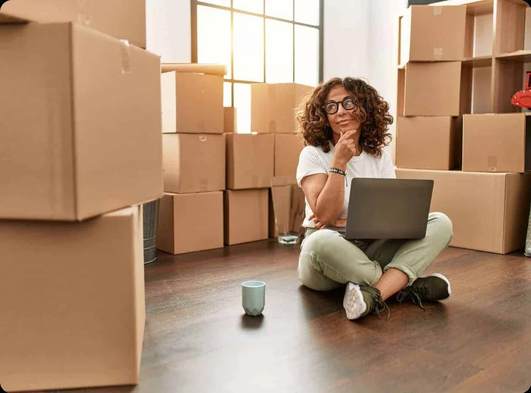 a woman sits on the floor with a laptop and coffee mug. She is surrounded by moving boxes and look contemplative