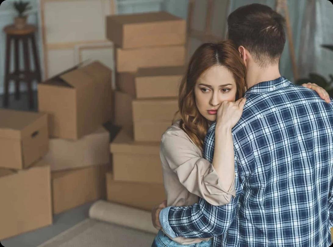 a couple comforts each other with an embrace. In the background, there are several stacked moving boxes