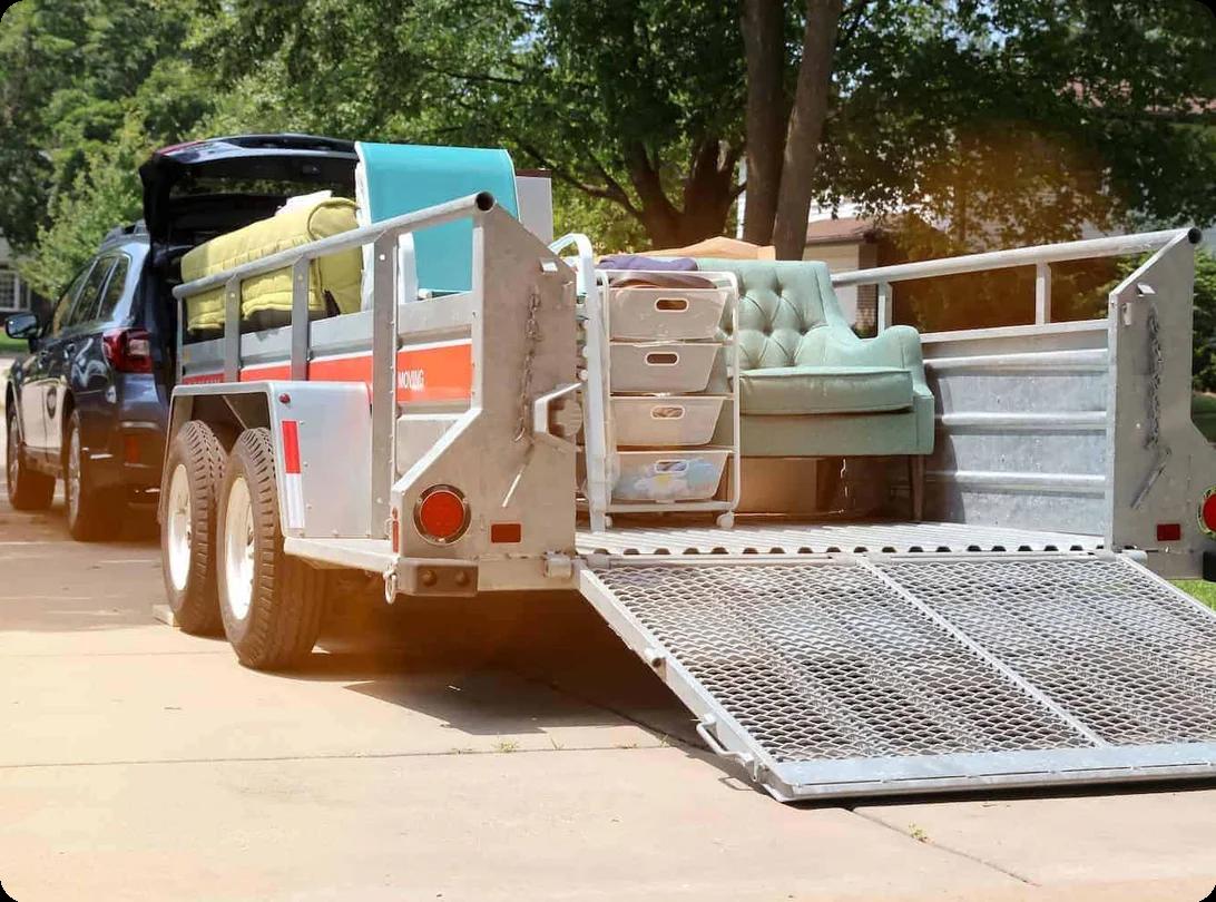 a car with a moving trailer hitched to it sits in a driveway. the trailer is full of furniture.