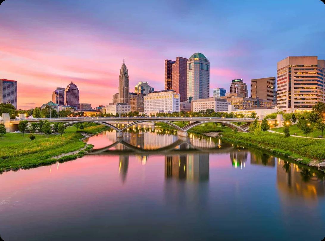 A view of downtown Columbus, OH at sunset. The buildings and skyscrapers border a river and bridge.