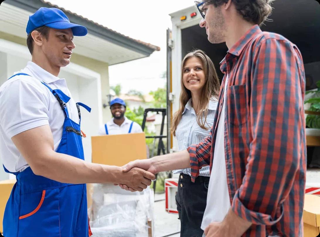 A couple shakes the hand of one mover while others move boxes out of a truck in the background