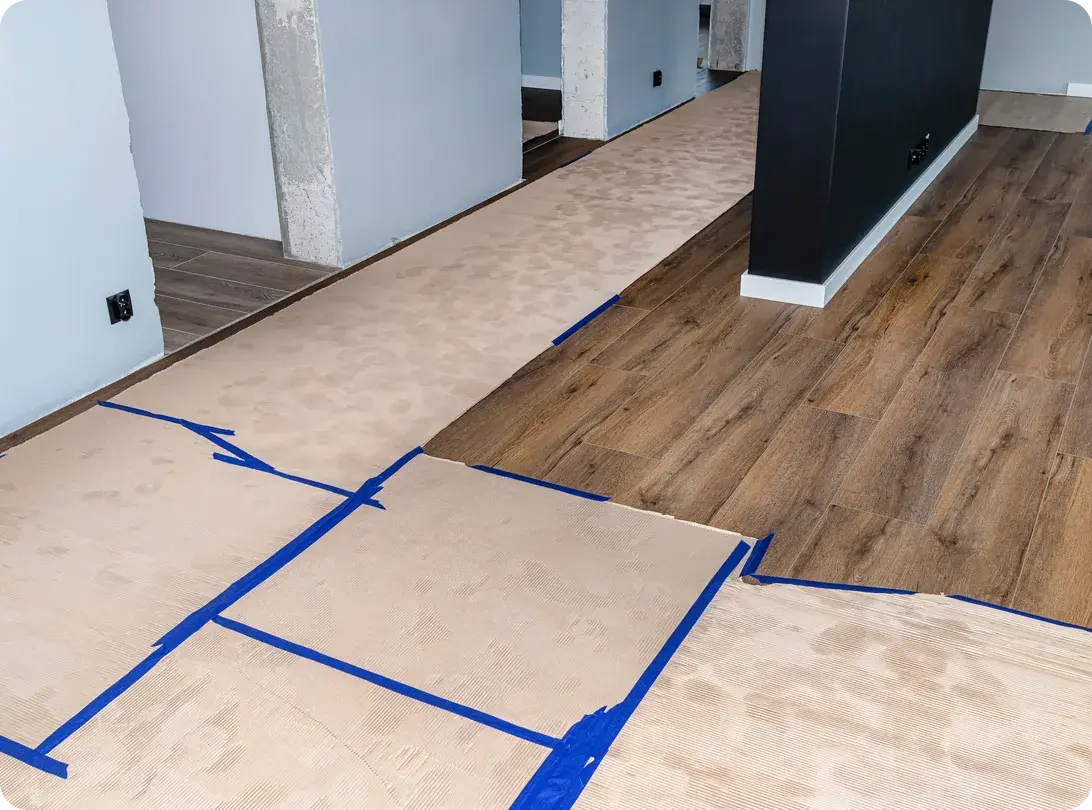 Sheets of corrugated cardboard floor protection taped down with blue painter's tape across a wood-look floor in an empty room