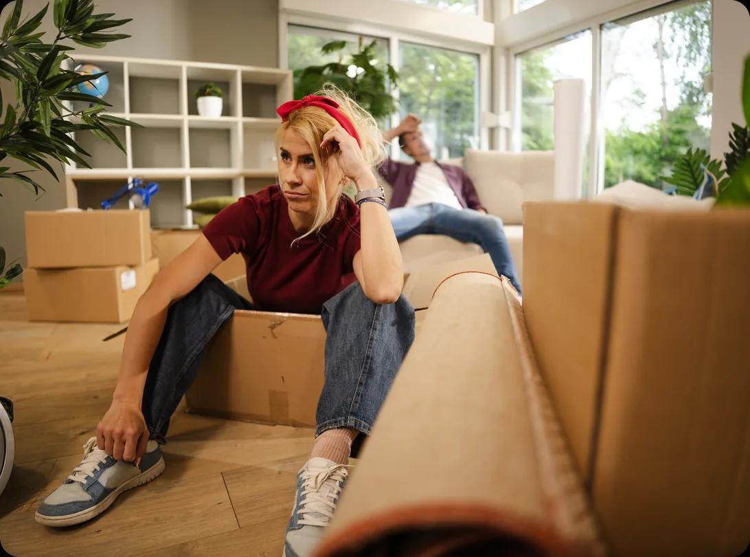 Someone sits on the floor of a bright living room surrounded by cardboard boxes and a rolled-up rug, resting their head on one hand, while another person sits on the sofa behind them.