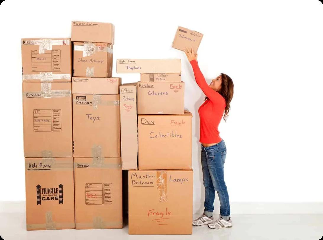 A woman reaches up to place a box at the top of a large stack of them, all labeled with various rooms and categories