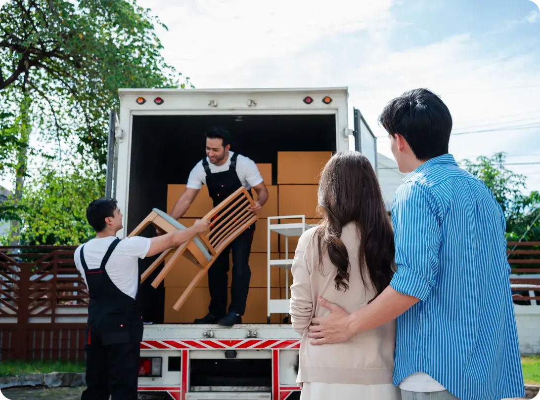 An Asian couple checks boxes while two uniformed movers unload furniture from a pickup truck at a new house
