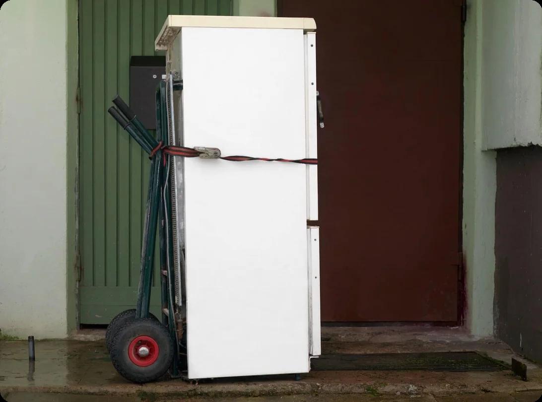 A fridge strapped upright to a dolly sits in a hallway