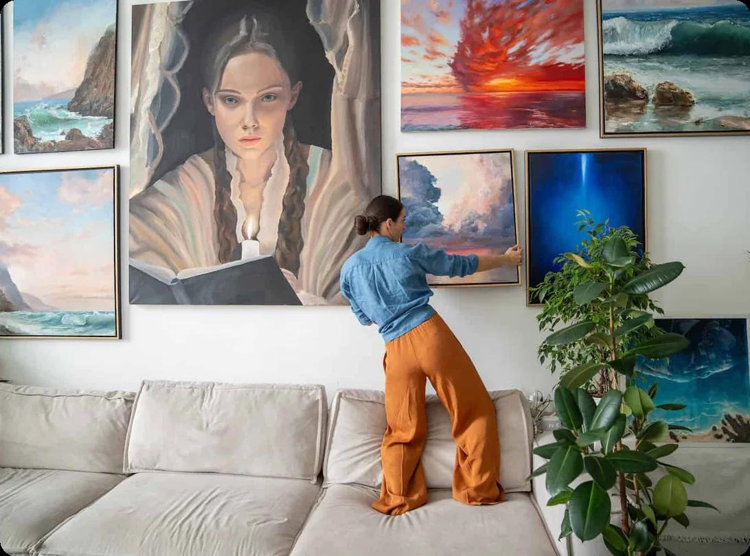 a woman takes down an oil painting from a tall wall that has many more covering its surface
