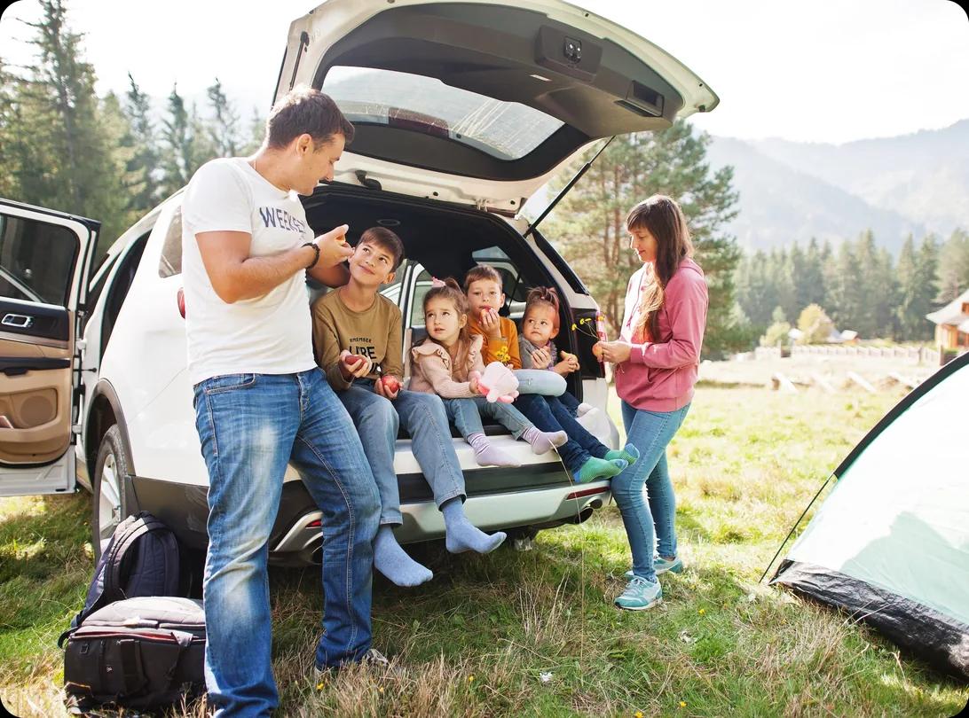 An adult and five children gathered at the open rear hatch of a white SUV parked on grass, four of the children sitting in the cargo area, with pine trees and mountains behind them