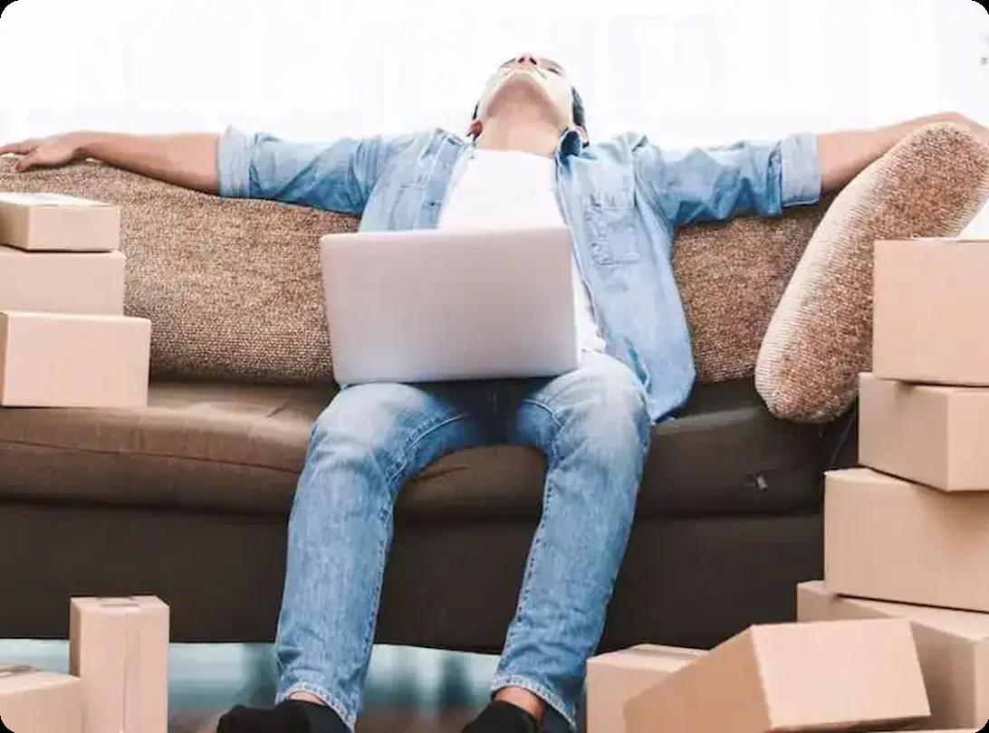 Man overwhelmed by moving stress, surrounded by cardboard boxes and laptop on couch