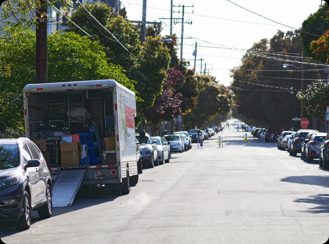 A moving truck parked at the curb with its loading ramp down on a tree-lined residential street, with cars parked bumper to bumper along both sides