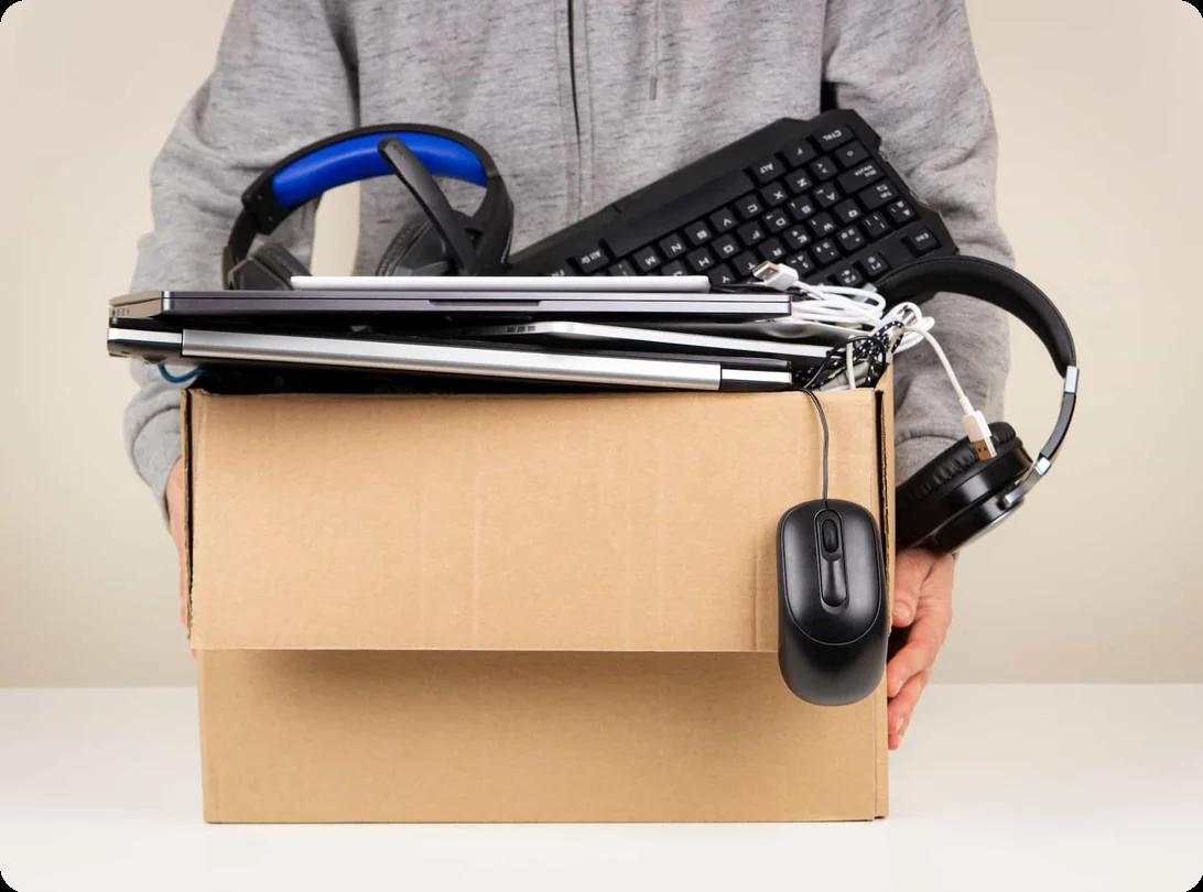 a person holds a box full of electronic waste, which include laptops, a keyboard, headphones, and a computer mouse