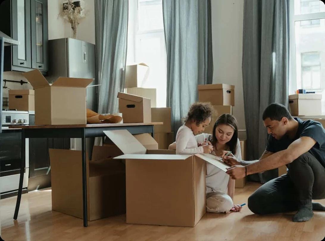 A man, a woman, and a child are seated on the living room floor, with packing boxes scattered around them, and they are drawing on the lid of the box