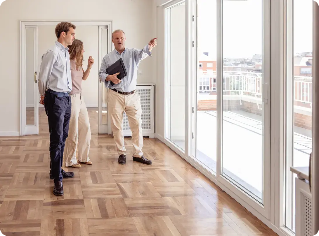 A real estate agent points out the view to a young couple touring an empty apartment with parquet floors and floor-to-ceiling windows
