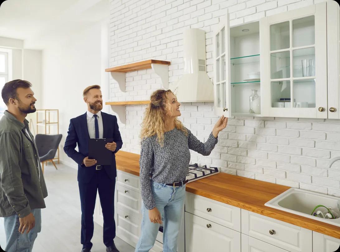 A couple tours an apartment kitchen with a letting agent, opening a glass-front cabinet to check the storage while the agent takes notes on a clipboard