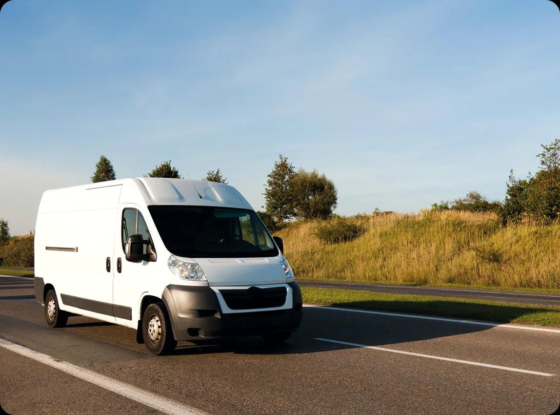 A white high-roof cargo van driving along a two-lane rural highway, with dry grass, scrub and trees on the verge under a clear blue sky
