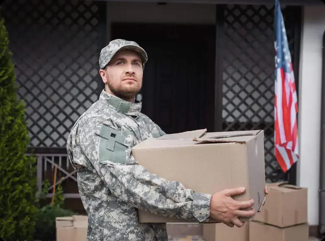 Soldier holding a moving box in front of a house with an American flag.