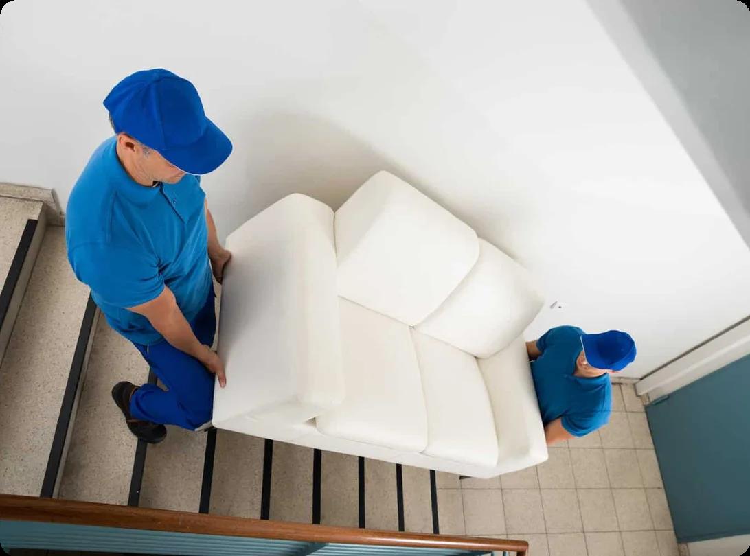 two movers in blue shirts and baseball caps move a white couch down a set of stairs