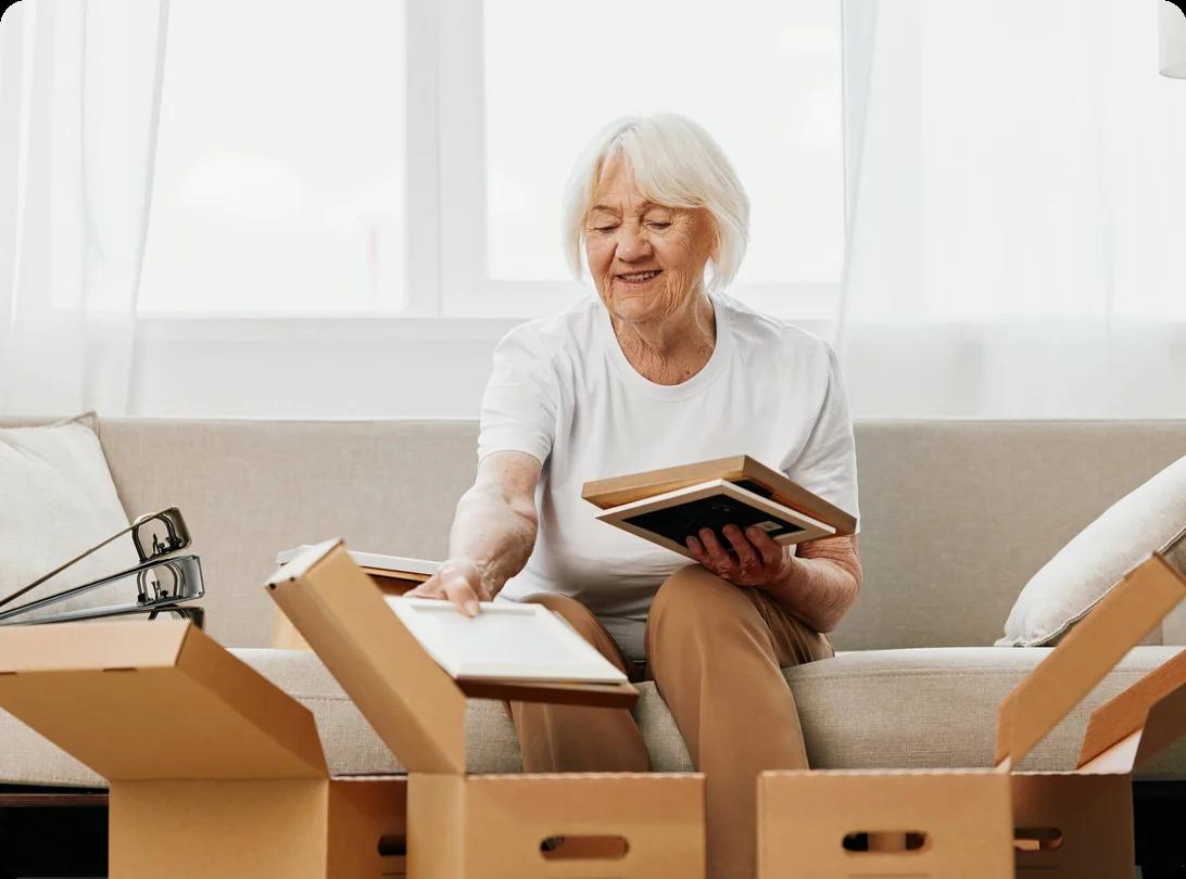 Elderly woman sits on a sofa at home with boxes collecting things with memories albums with photos and photo frames moving to a new place cleaning things and a happy smile.