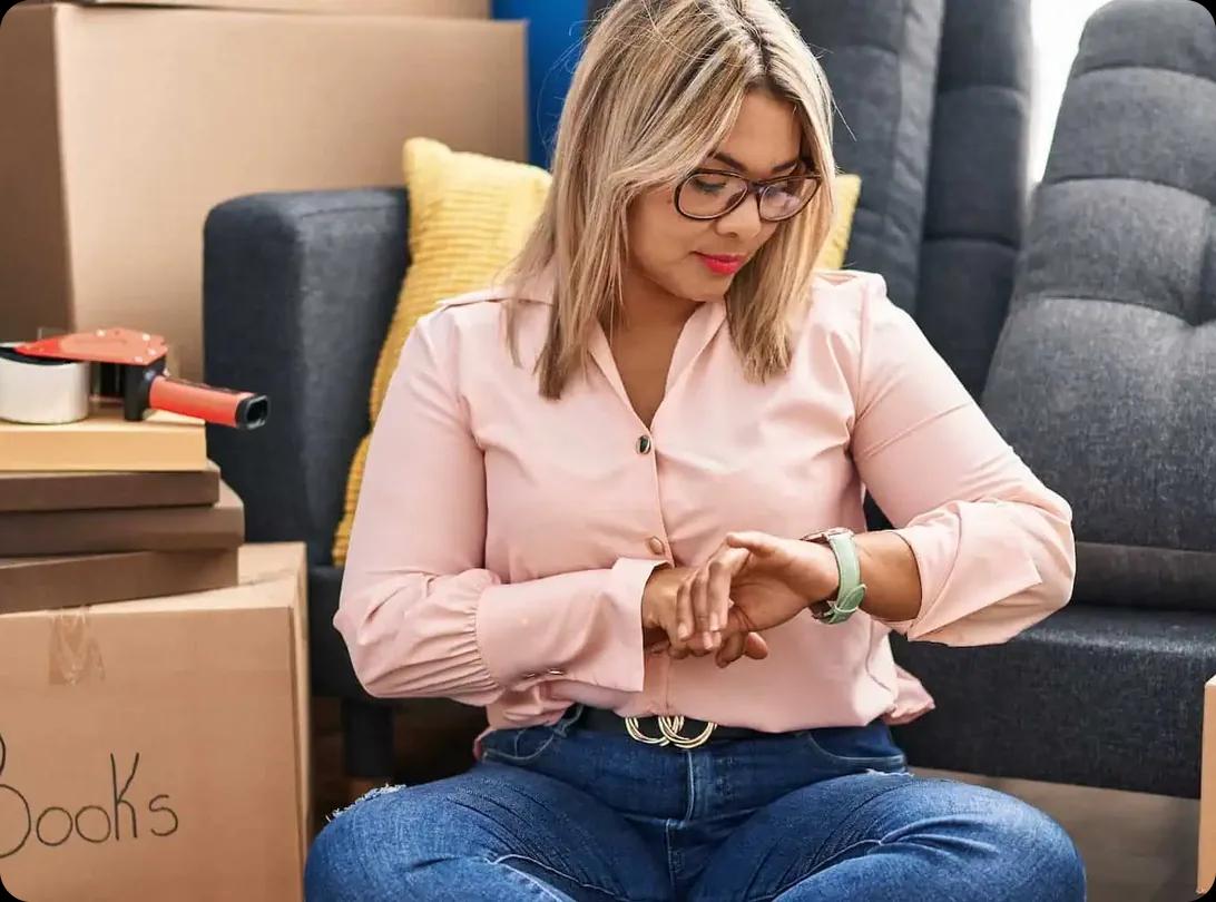 A woman sitting among packed moving boxes checking her watch.