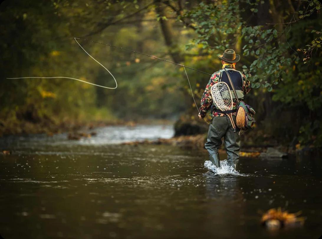 A man knee deep in a river. He's wearing waders, tackle, and fishing gear, and is in the middle of a cast.