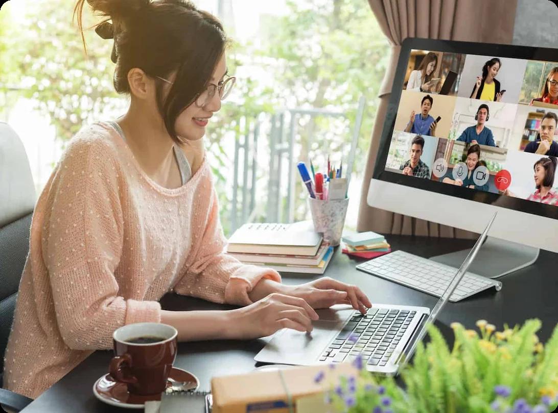 A woman works from home at a laptop on a spacious desk while a virtual meeting appears on another monitor