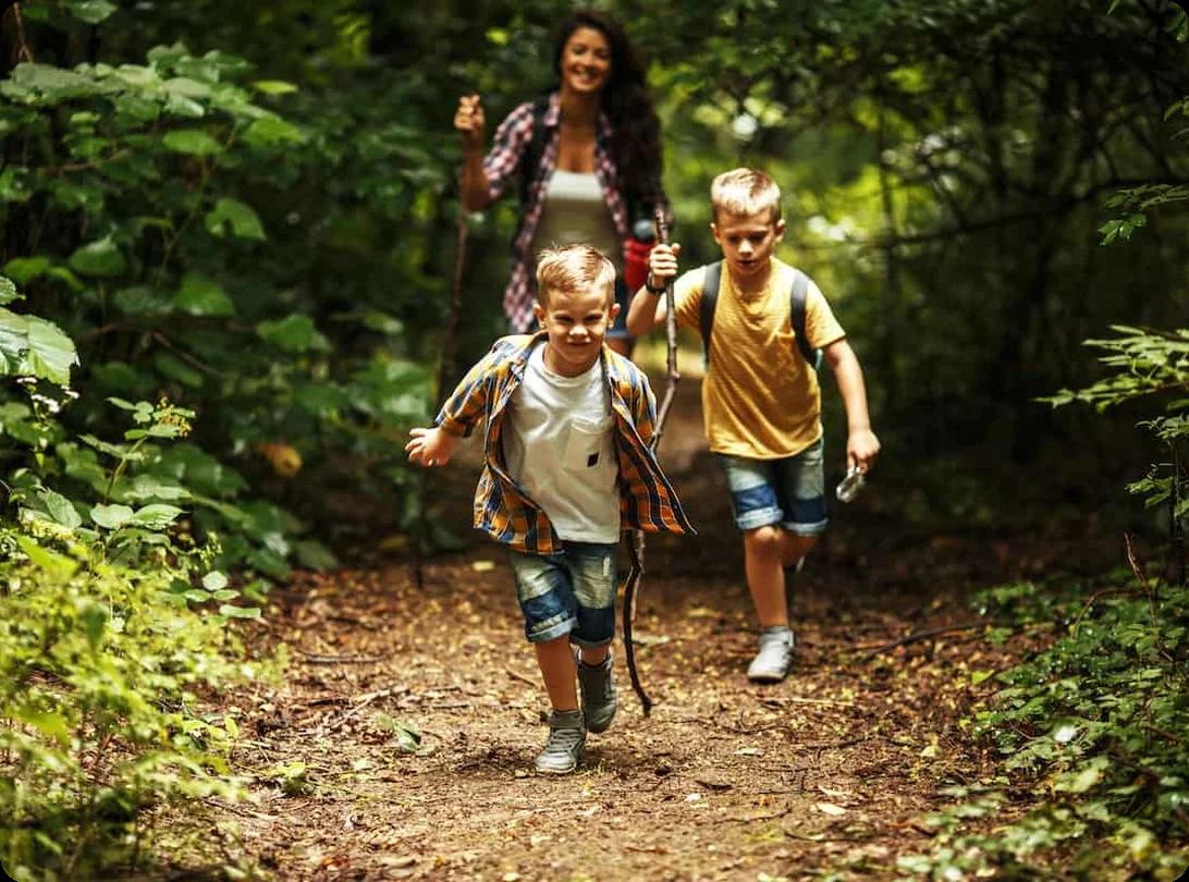 Two children exploring a forested hiking trail with backpacks on a sunny day