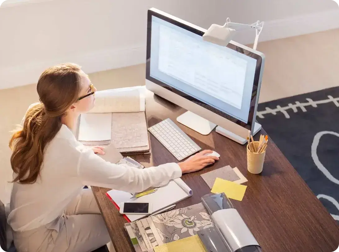 A woman working at a home office desk with a monitor, keyboard, notebooks and paperwork.