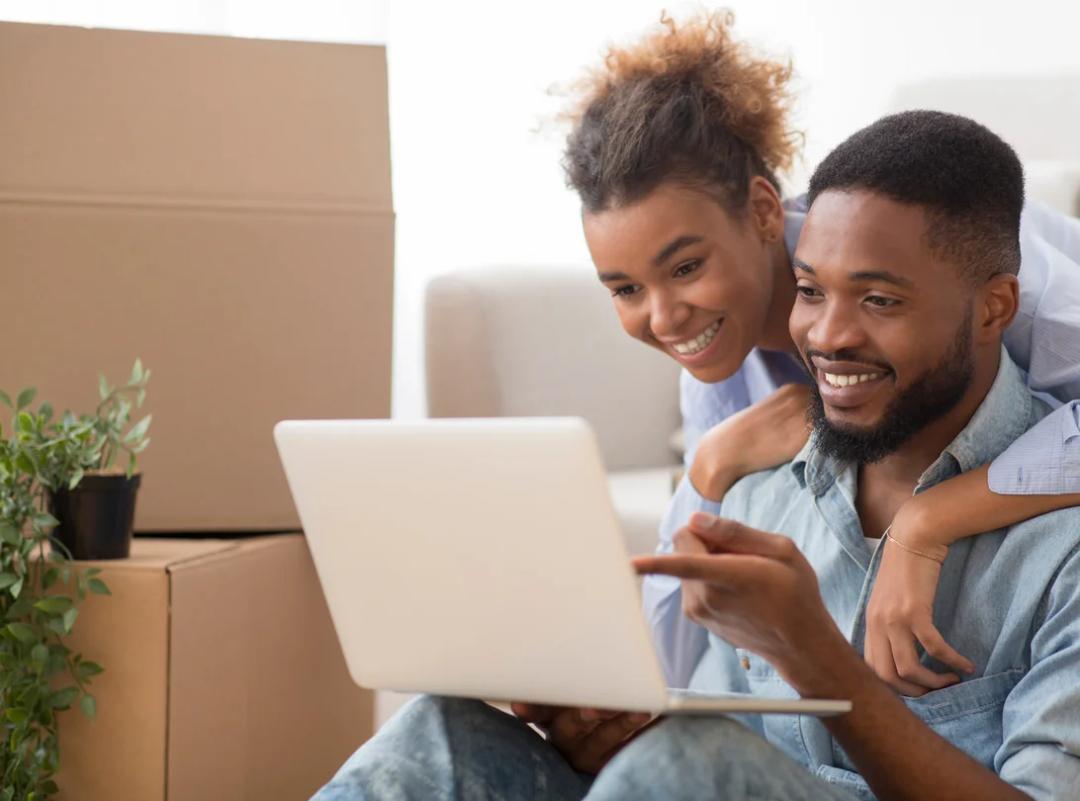 A smiling couple using computer surrounded by moving boxes