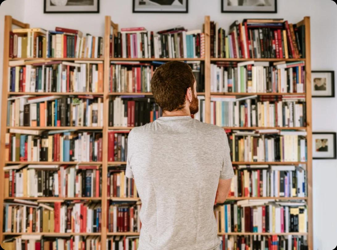 a man stands contemplatively in front of a full bookshelf that takes up one wall of a room