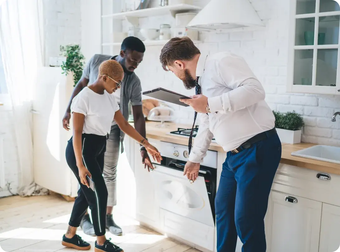 A young couple looks at the oven in a bright apartment kitchen while an agent holding a tablet shows them the unit