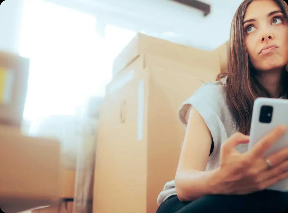 A woman sitting among packed moving boxes looking uncertain while holding her phone.