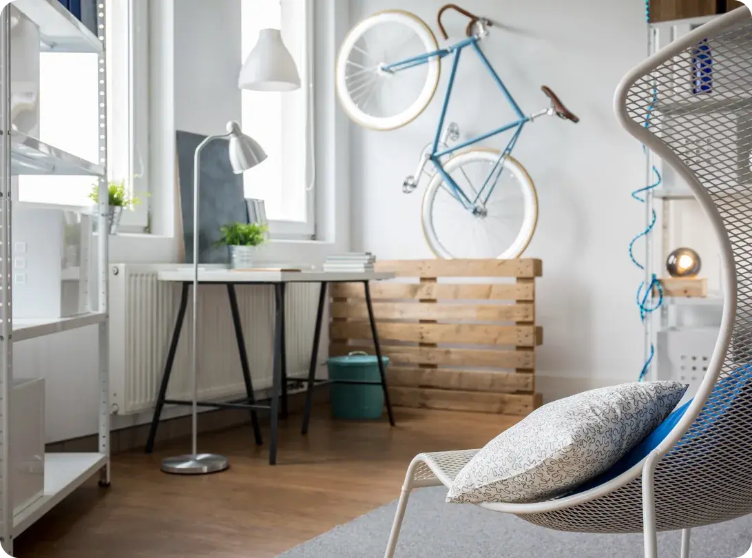 A small bright apartment room with a bicycle mounted on the wall above a wooden pallet divider, open shelving, a slim trestle desk and a mesh chair