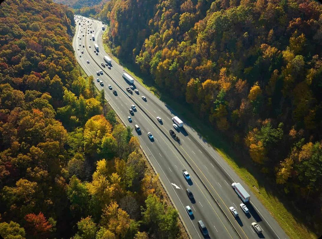 an array of cars and trucks are driving in both directions down a long highway in the middle of a very wooded area