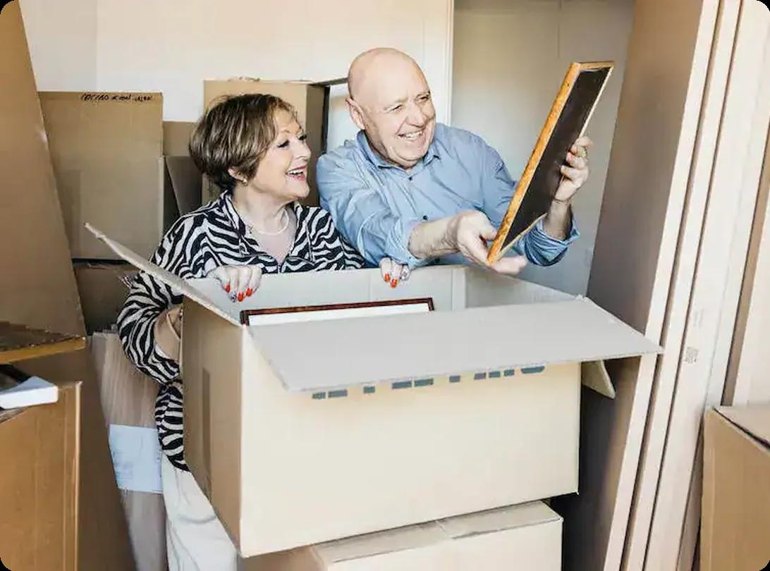 Older couple sorting framed photos and keepsakes while packing boxes during a downsizing move
