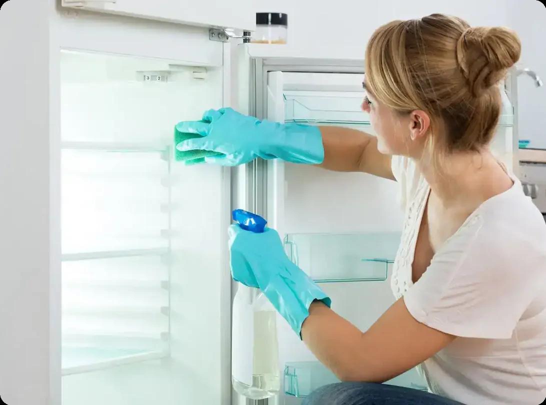 Woman cleaning fridge interior with gloves and spray bottle