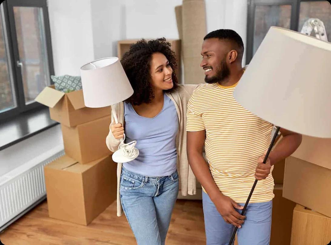 A couple carrying lamps in a room filled with boxes smile at each other