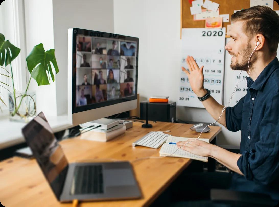 A man wearing earbuds waves at a grid of colleagues on a video call displayed on a desktop monitor, seated at a wooden home-office desk with an open laptop, a notebook and a wall calendar behind him