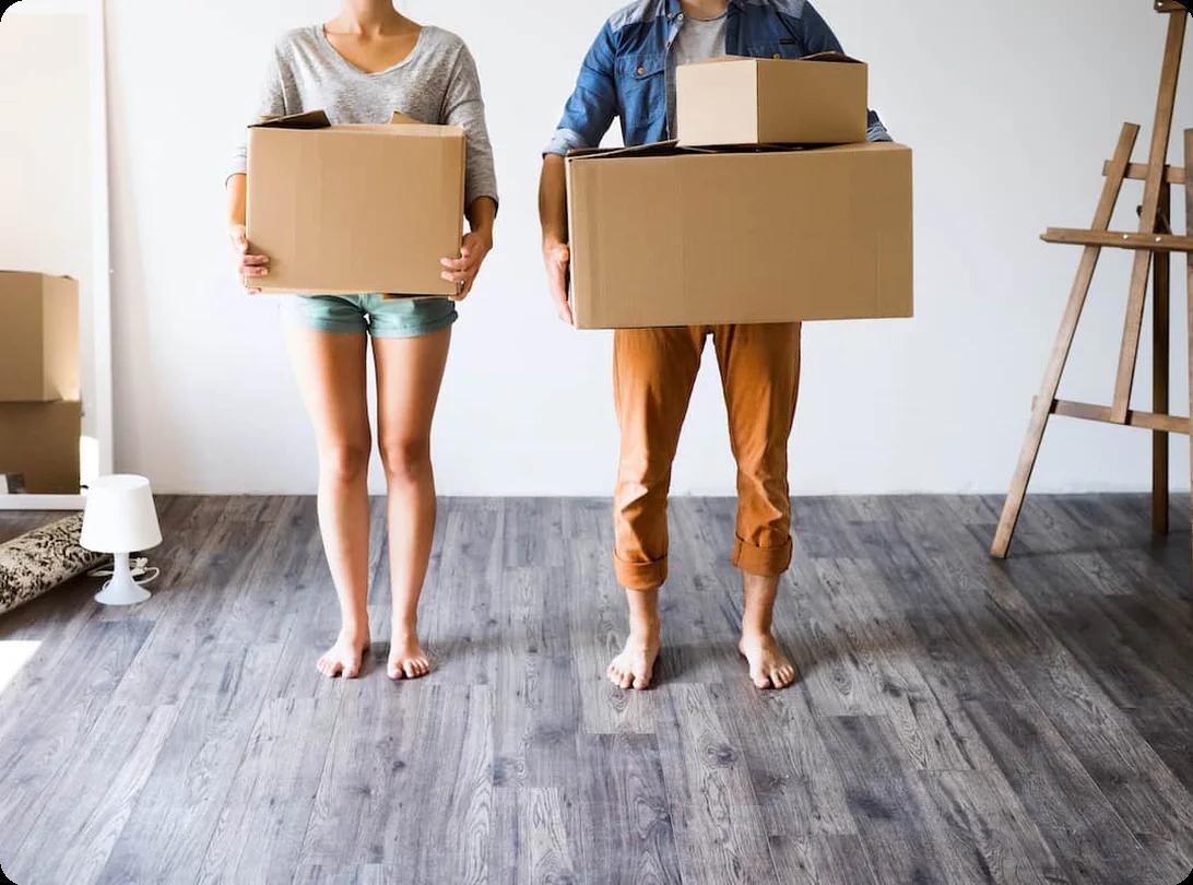 A young couple holding cardboard moving boxes in an empty room with hardwood floors, ready for an out-of-state move