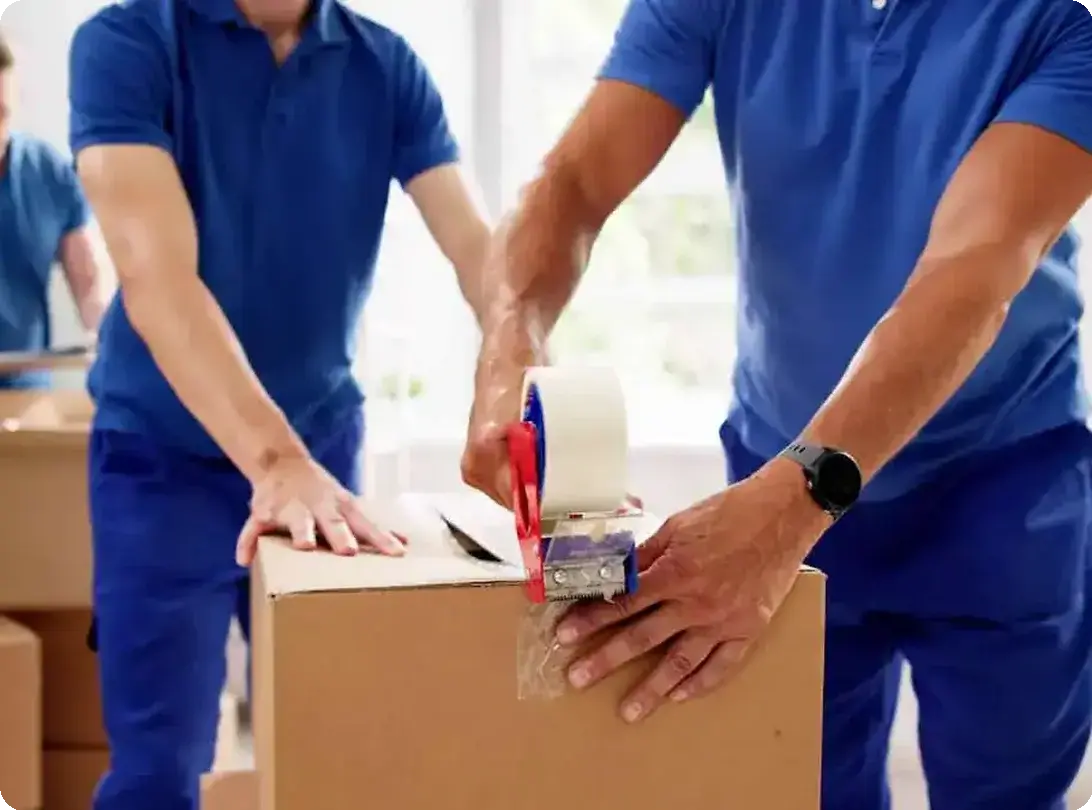 Professional movers sealing a cardboard box with packing tape while preparing items for a move