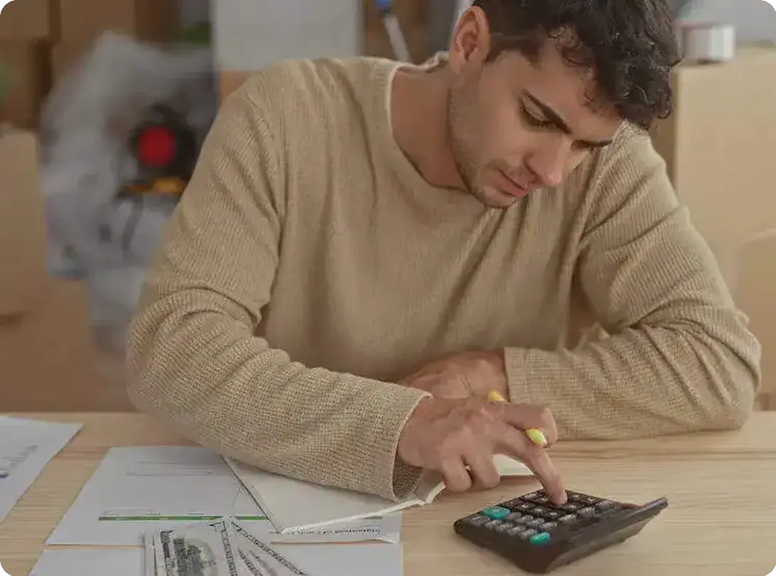 A man calculating moving costs with calculator, paperwork, and packing boxes at the back