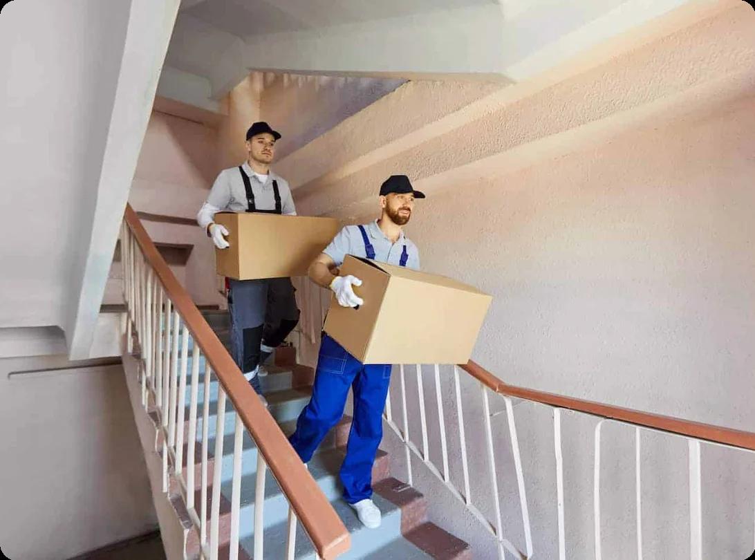 Movers in blue overalls carry boxes down a set of stairs in an apartment building