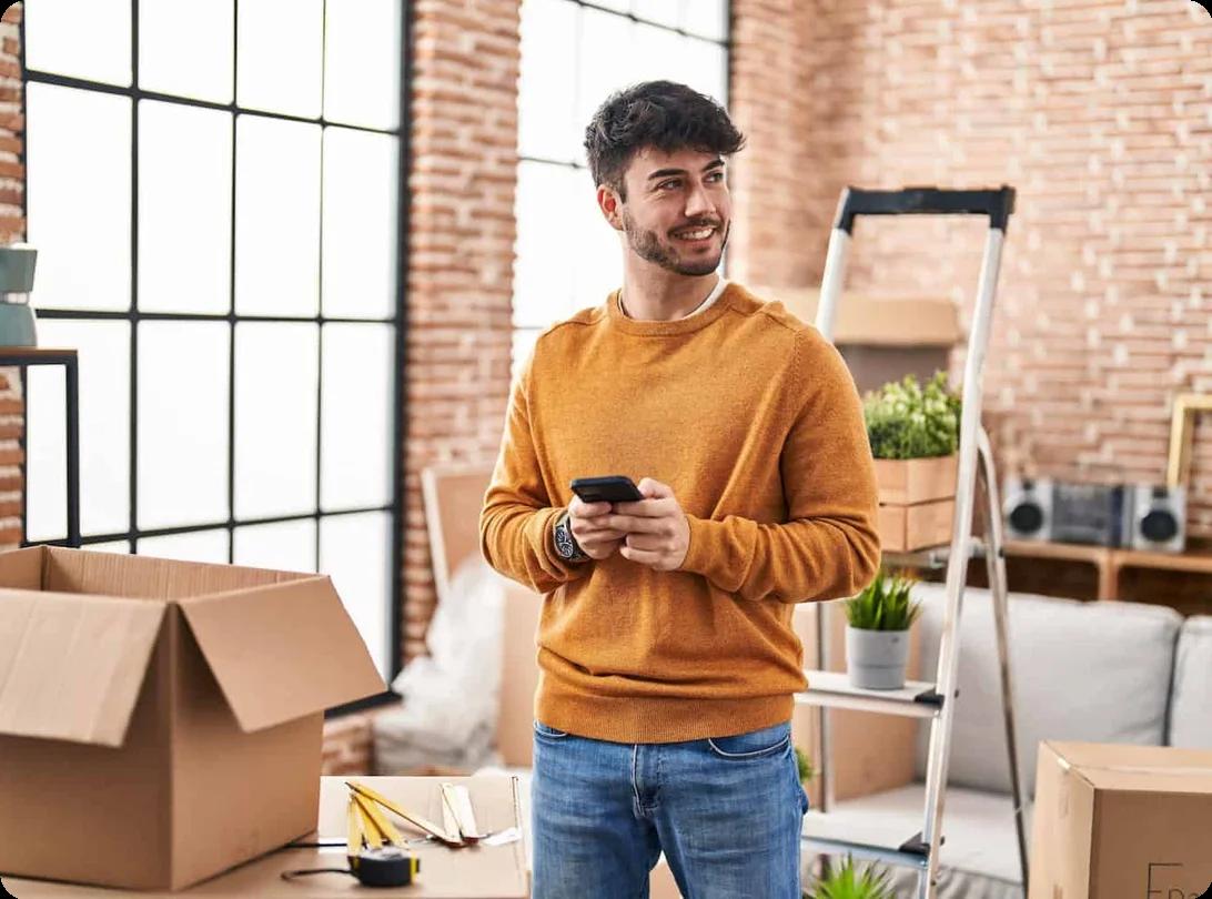 a man stands in a room full of moving boxes while holding his smart phone