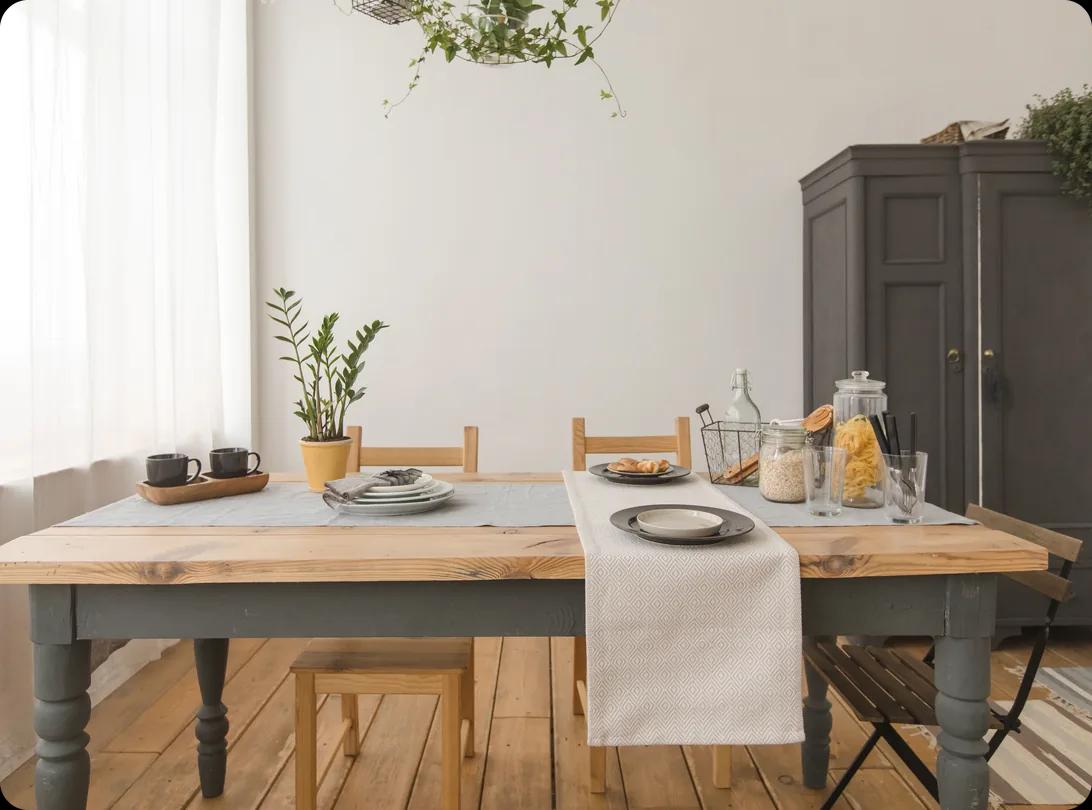 A wooden dining room table with a natural pine top and grey-painted legs, set with plates, mugs and a linen runner in a bright room with wood floors.