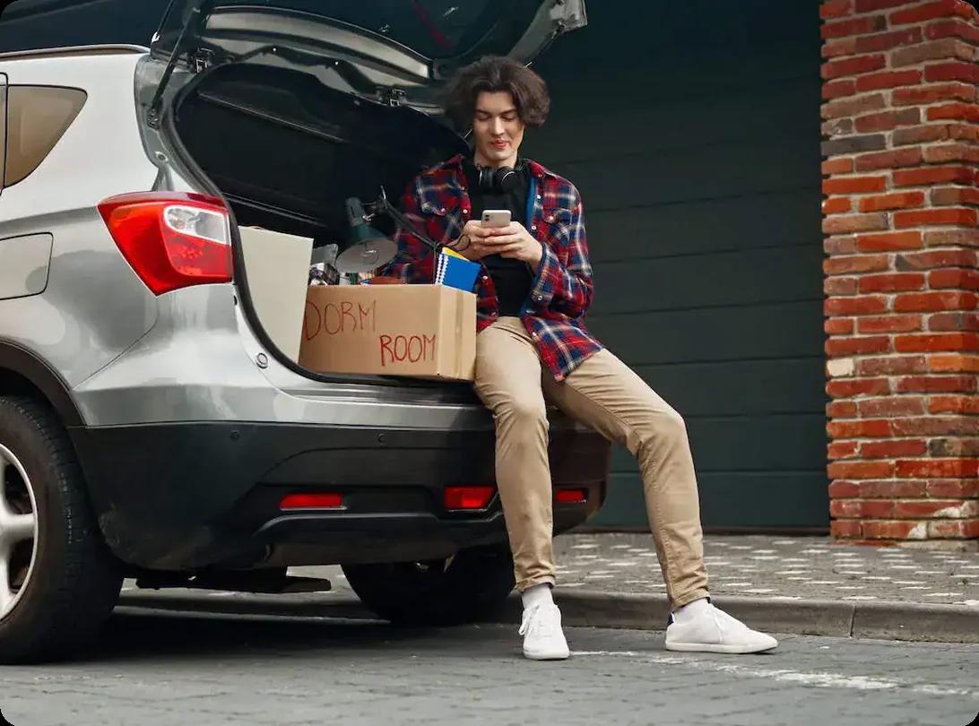 an image of a young man sitting in the open trunk of an SUV on his phone. There are moving boxes visible sitting next to him.