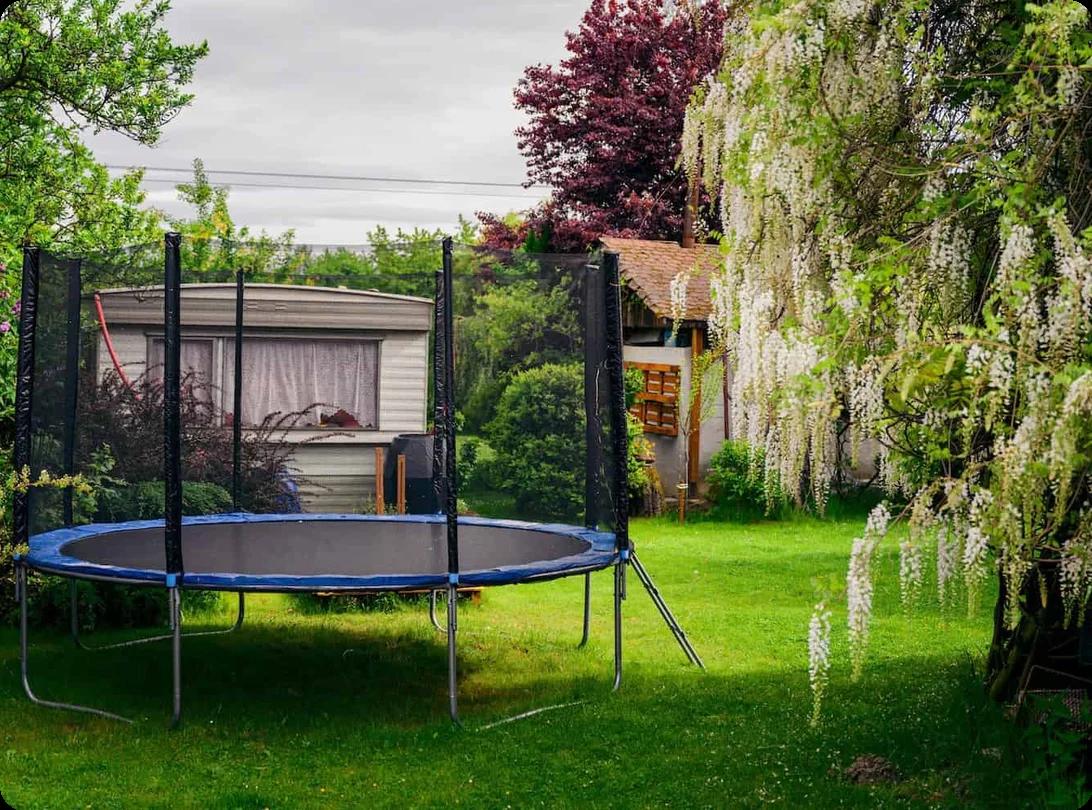 A trampoline with a net sits in the corner of a backyard with a green lawn and trees