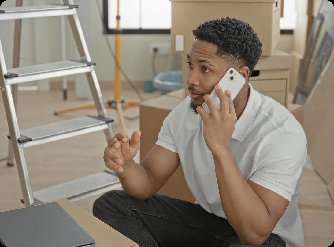 A man sitting on the floor among stacked moving boxes and a step ladder, talking on a mobile phone with one hand raised mid-gesture