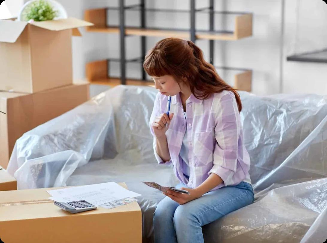 A person at a kitchen table with a budget worksheet, calculator, and moving boxes planning a low-cost move