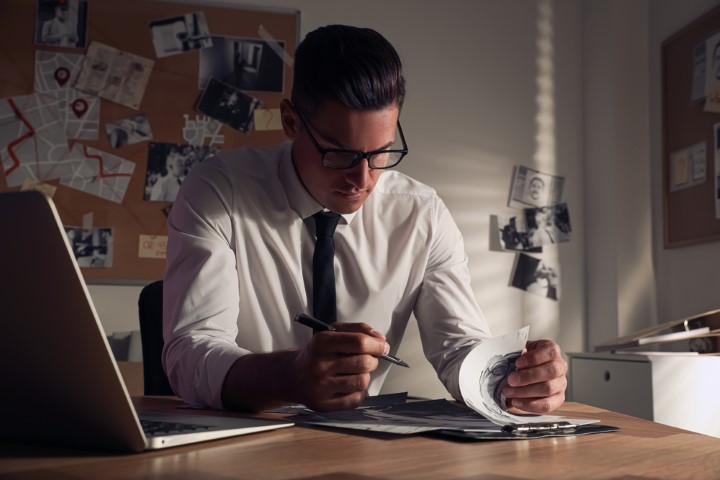 Man with glasses sitting at a desk holding a pen looking at clipboard